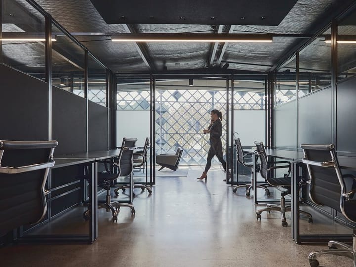 Dark open-plan workspace with glass partition offices, exposed ceiling and geometric window beyond
