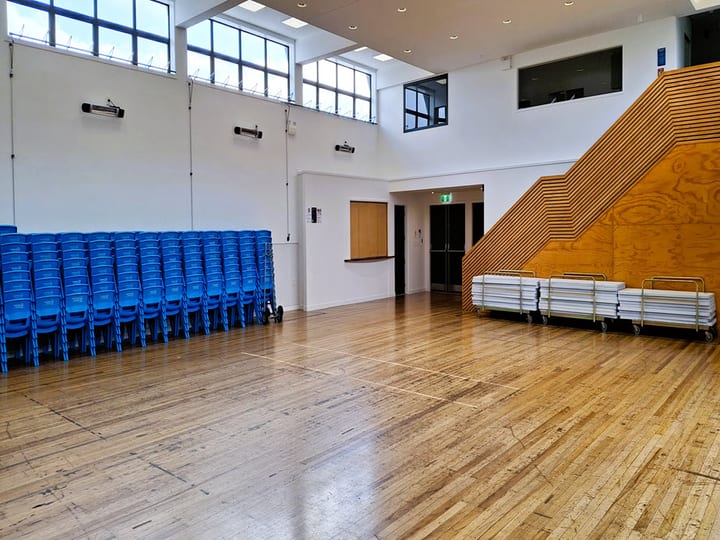 Open hall space with timber floor, stacked blue chairs and trestle tables on trolleys, timber stair wall visible