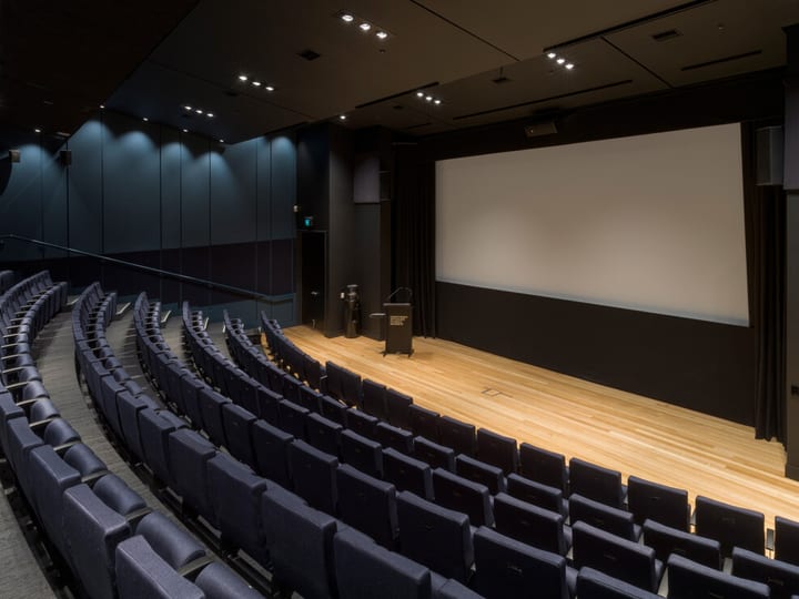Dark blue-walled auditorium with curved rows of blue upholstered seats facing a timber-floored platform with projection screen