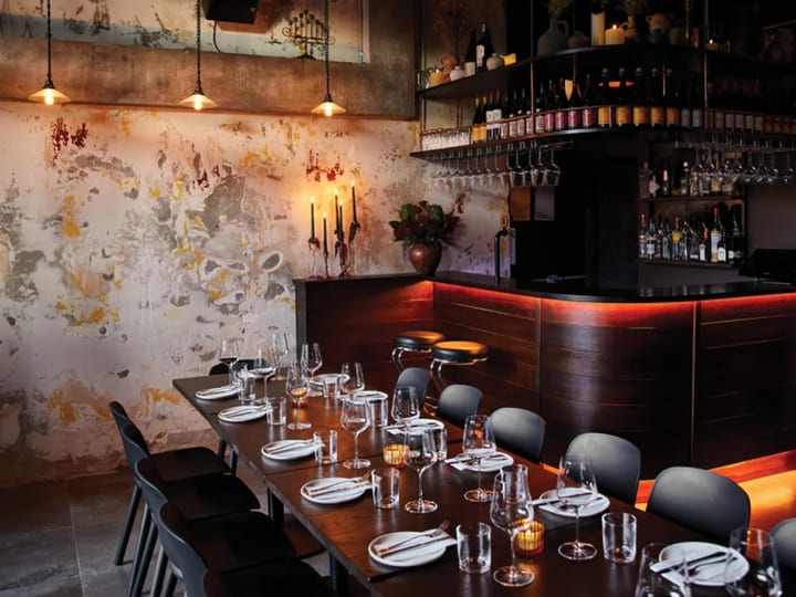 Dark dining room with distressed plaster wall, warm-lit timber bar, pendant lights, and set dining table