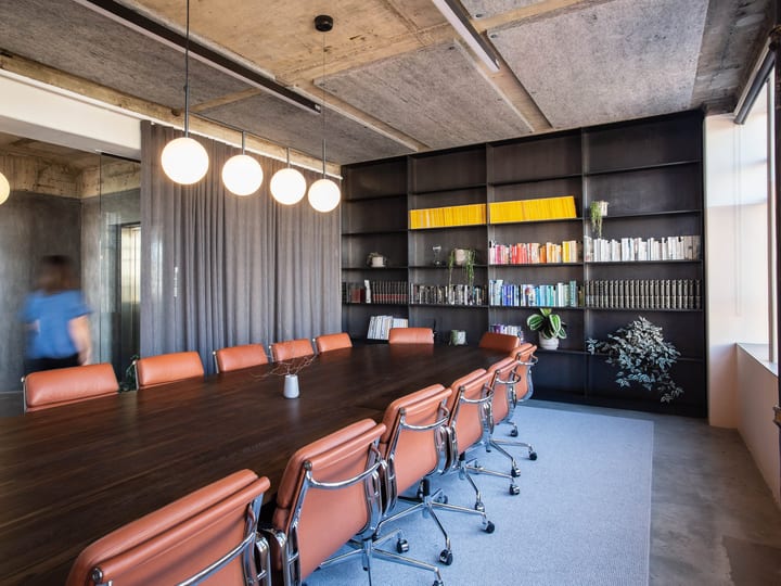 Exposed concrete ceiling boardroom with dark shelving wall, tan leather chairs, dark timber table, and globe pendant lights