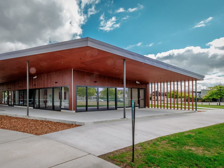 Low flat-roofed timber building with a wide canopy, glass frontage, and paved surrounds on open grounds