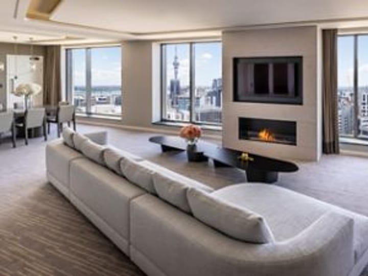 Bright suite lounge with large grey sectional sofa, linear fireplace, floor-to-ceiling windows overlooking city and harbour