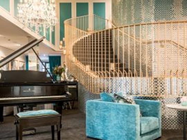 Ornate hotel lobby with grand piano, curved staircase, crystal chandelier, and teal velvet armchair