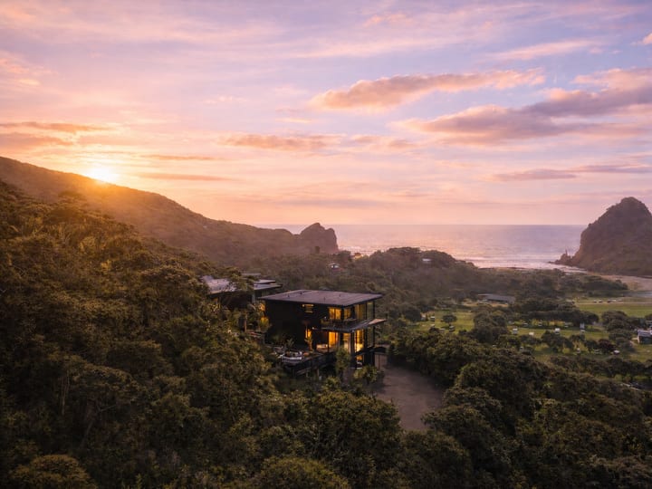 Dark multi-level building above treeline with ocean and rock stack visible at sunset