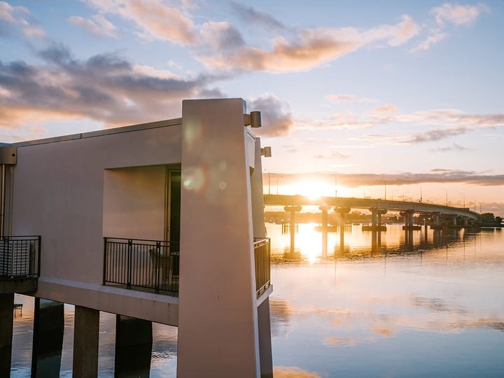 White multi-storey building on stilts over water with jetty and trees behind