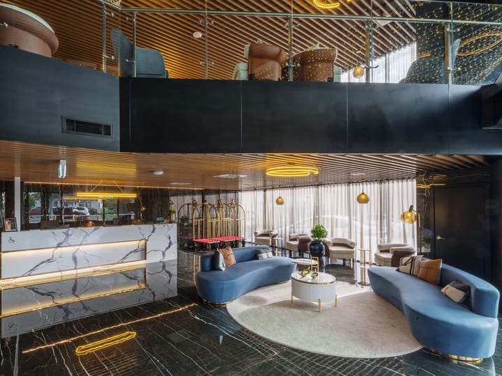 Dark marble hotel lobby with curved blue sofas, mezzanine above, timber slatted ceiling, and gold pendant lights
