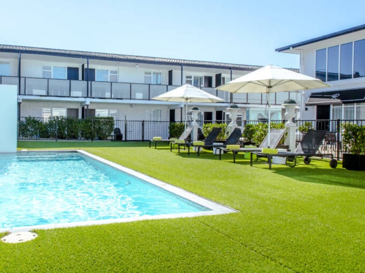 White two-storey hotel building with pool, sun loungers, umbrellas, and manicured lawn
