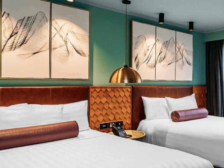 Twin hotel room with teal walls, leather headboard, bold abstract artwork, and copper pendant light