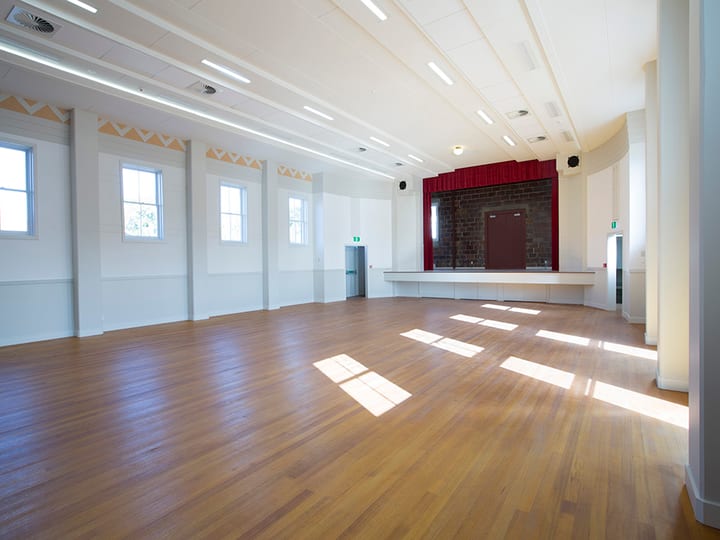 Open hall with timber floor, tall windows, and a raised platform with red curtain at far end