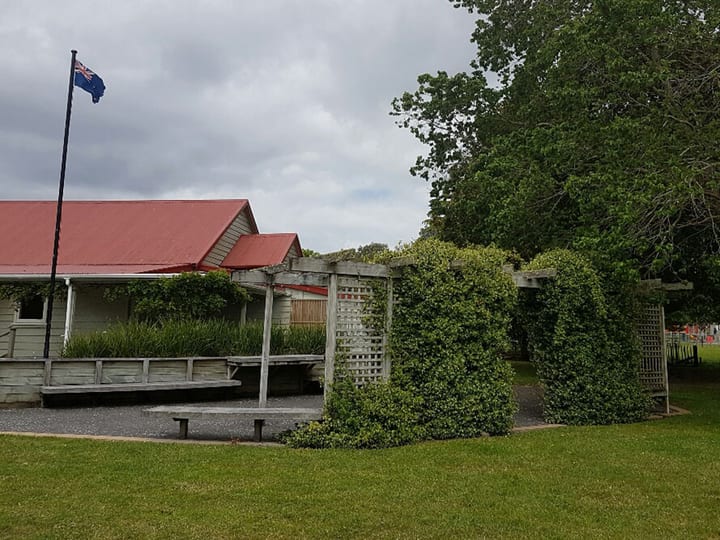 White weatherboard hall with red roof, flagpole, and clipped hedge archway