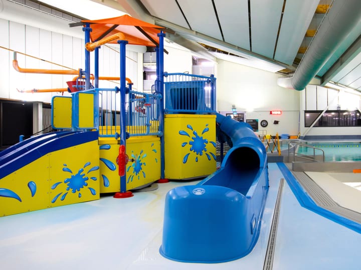 Indoor water play area with coloured slide and play structures beside a pool