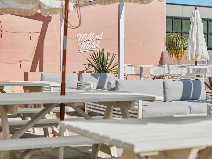 Outdoor seating area with fringe umbrellas, bench tables and neon sign on pink wall