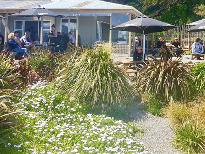 An outdoor seating area with dark umbrellas and picnic tables among native flax beside the hotel
