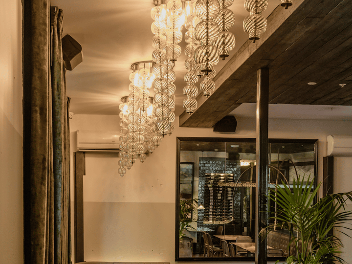 Warm dining room with globe chandeliers, timber ceiling, banquette seating, and wine display beyond glass partition