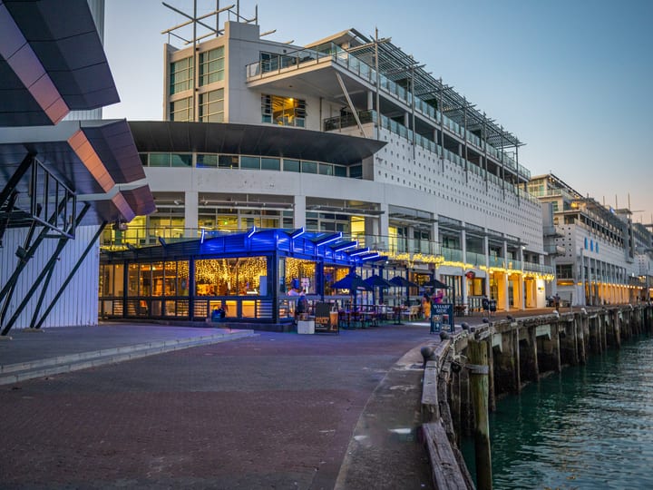 Waterfront bar and dining pavilion with blue neon canopy, dusk, harbour backdrop