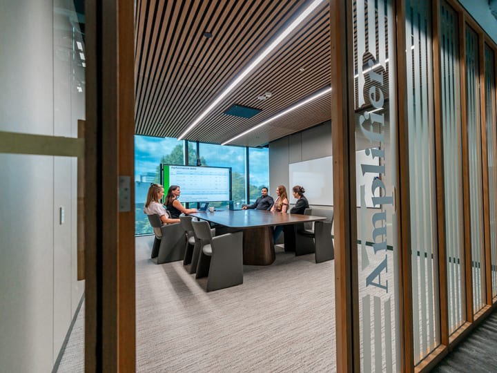 Glazed meeting room with a round dark table, people seated, and timber louvre screens outside