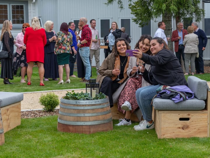 Guests gathered on a lawn beside a white weatherboard building with outdoor barrel furniture
