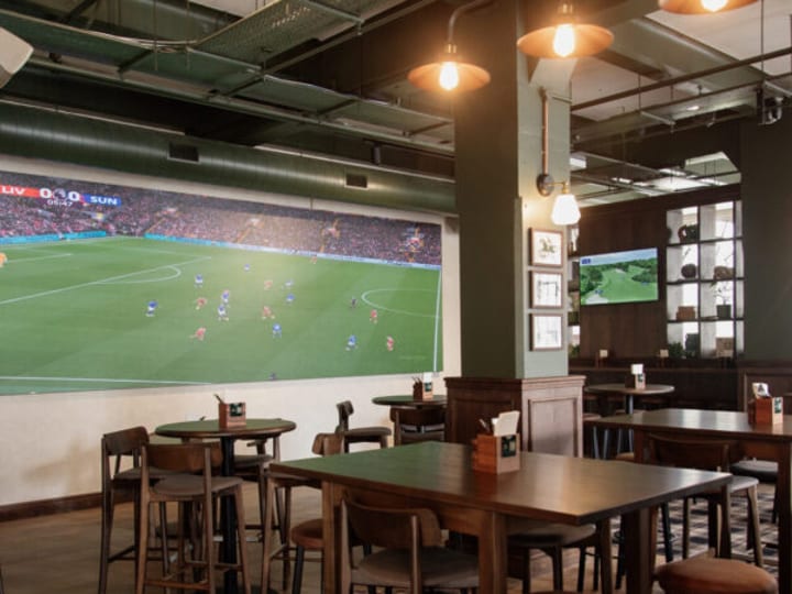 Dark green bar interior with large screen, plaid carpet, and high wooden tables