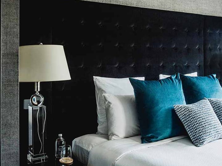 Hotel room with dark tufted headboard, teal cushions, and bedside lamp