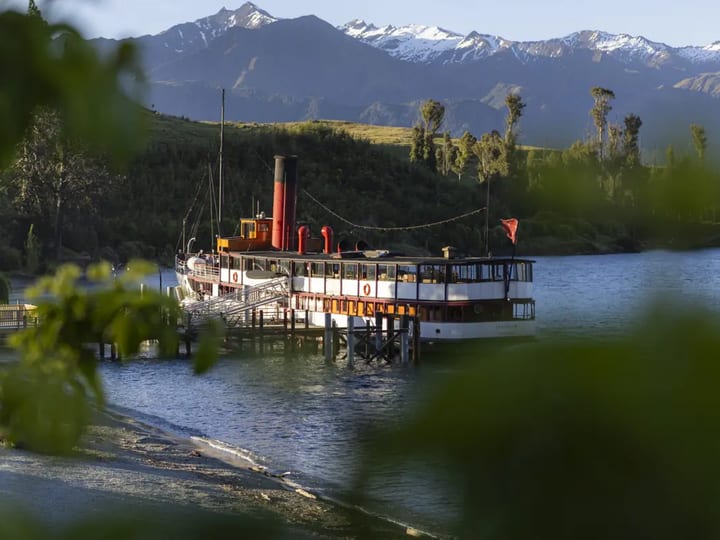 Vintage steamship moored at a timber wharf with trees and snow-capped mountains beyond