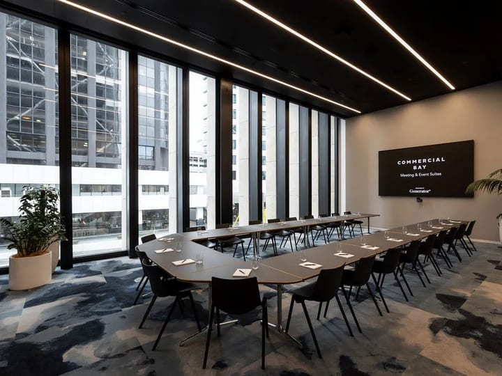 Modern boardroom with U-shaped table, dark chairs, floor-to-ceiling windows with city high-rise views and wall-mounted screen