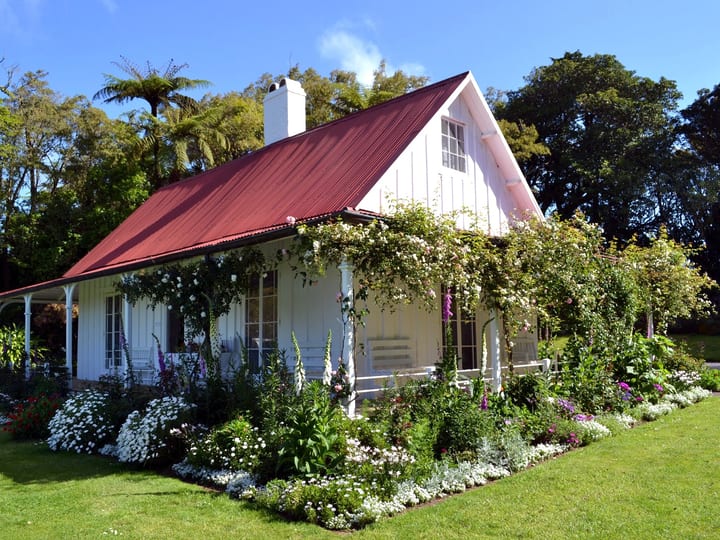 White cottage with red roof surrounded by cottage garden in full bloom