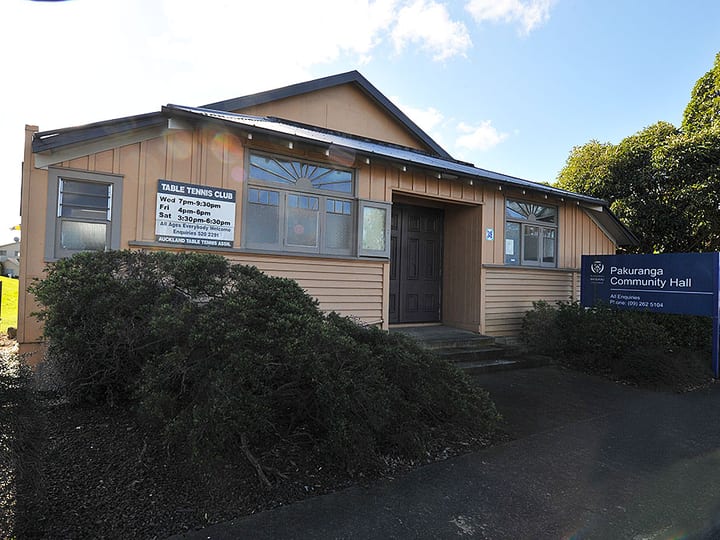 Timber-clad community hall with dark door, signage and overgrown shrubs