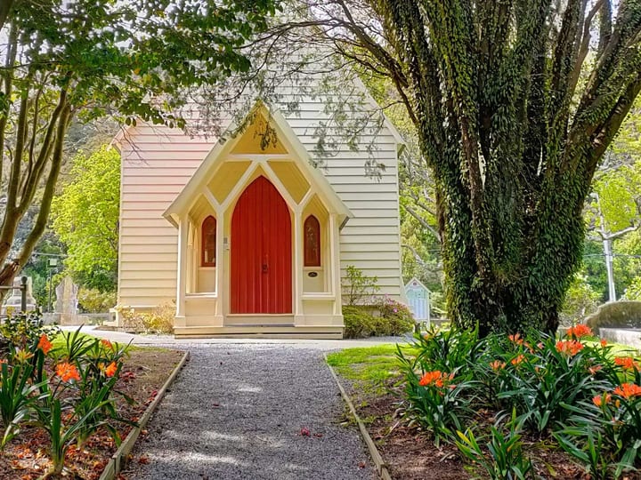Small cream timber church with red roof and gardens in foreground
