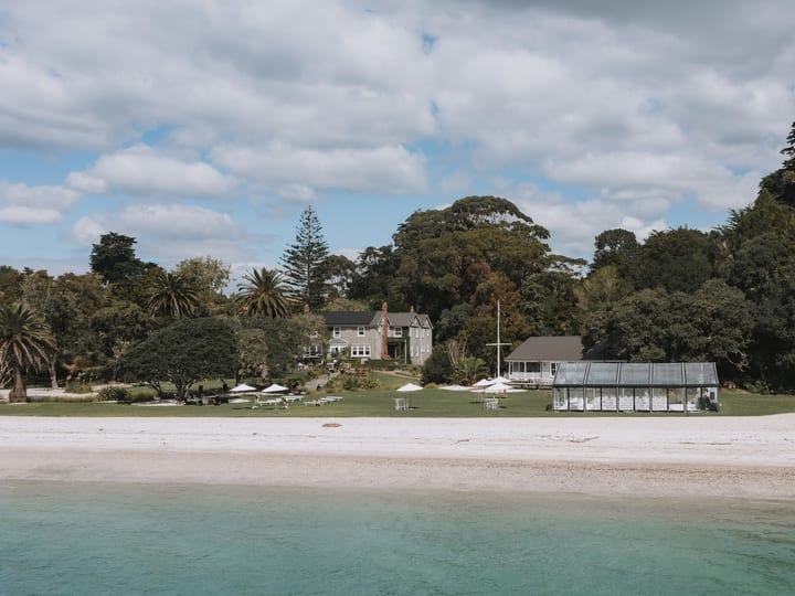 Stone homestead and glass marquee on lawn beside sandy beach with trees behind, viewed from water