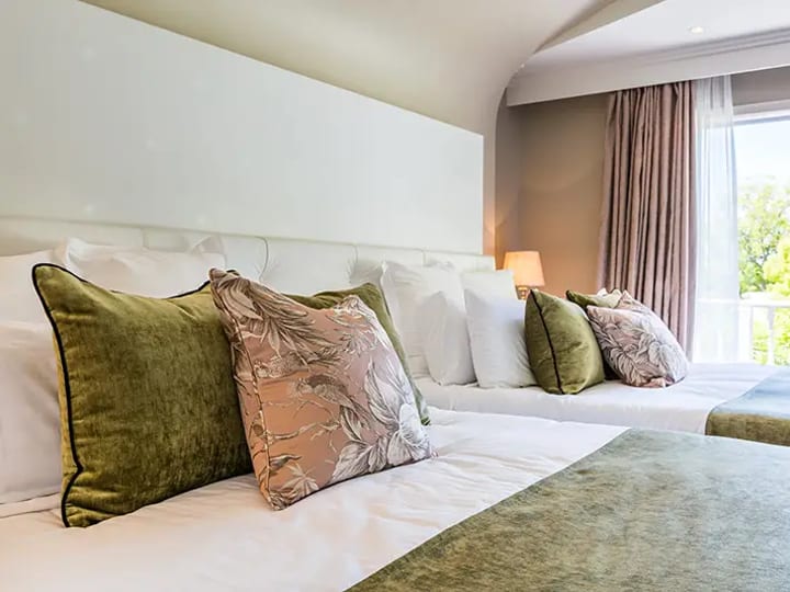 Twin hotel room with green and pink cushions, white bedding, balcony with tree view