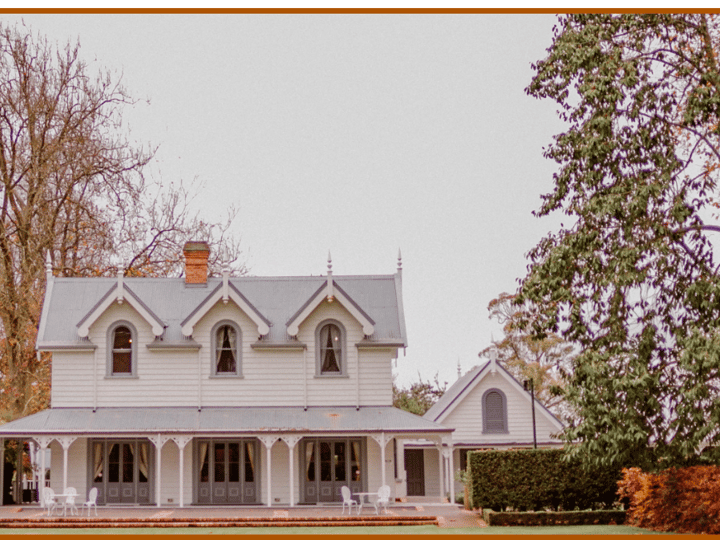 White Victorian-era homestead with steep roof, dormer windows and wrap-around veranda in formal hedged gardens with autumn trees