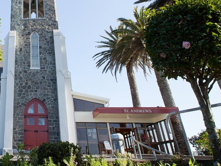 Dark stone church with tall bell tower, red arched doors, and trees lining the path