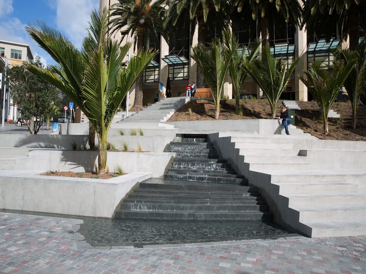 Water cascade over dark stone steps with trees and city buildings in background
