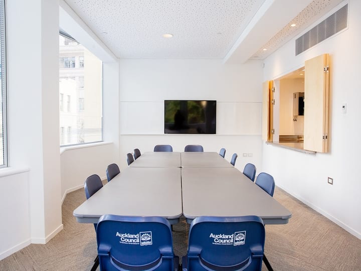 Meeting room with long table, blue chairs and wall-mounted screen beside a servery window