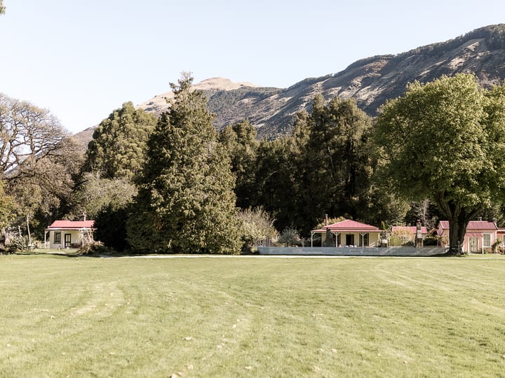 Red-roofed accommodation buildings across a wide flat lawn with mature trees and mountains behind
