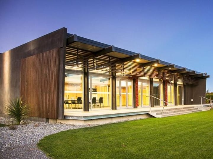 Low dark-clad building with timber louvred pergola, lit interior visible through full-height glazing, lawn foreground at dusk
