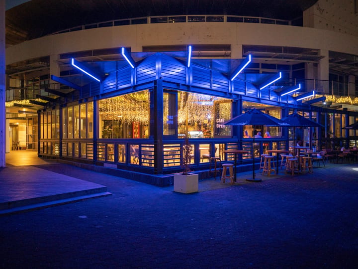 Glass-fronted venue with blue neon canopy, fairy lights inside, outdoor stools at night