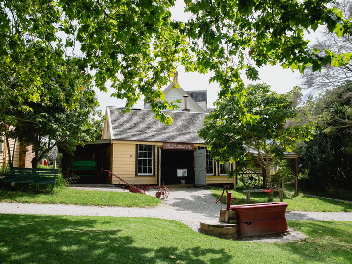Yellow weatherboard colonial buildings with gravel courtyard, trees and red farm implements