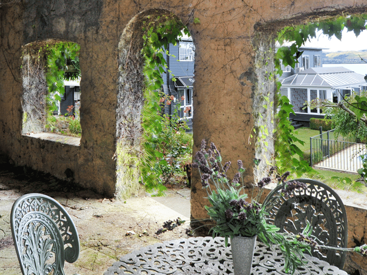 Weathered stone garden wall with open rectangular windows draped in climbing plants, cast-iron furniture on stone floor