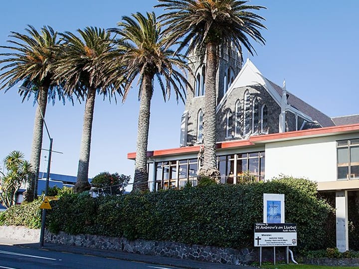 Stone church with attached white hall building, palm trees, and red cross on the facade