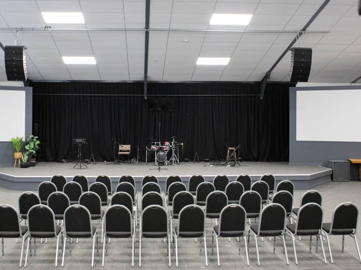 Modern church hall with rows of chairs facing a raised platform, flanked by large screens and a lighting rig