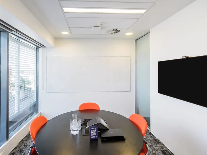 Small meeting room with round dark table, four orange chairs, whiteboard, wall-mounted screen, and window with louvre blind