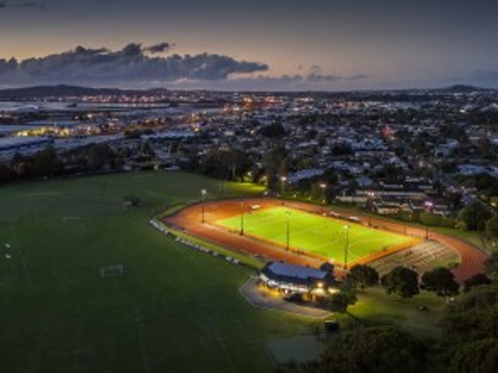 Illuminated sports field and running track at dusk with a city horizon in the background