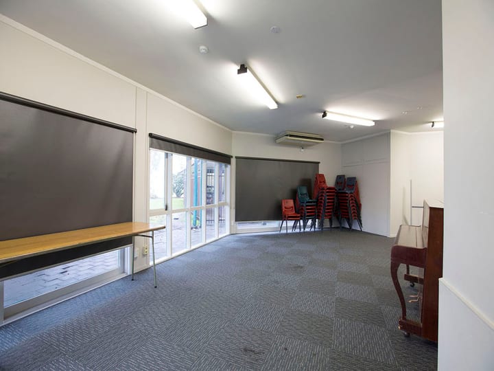 Plain carpeted room with stacked chairs, roller blinds, and upright piano