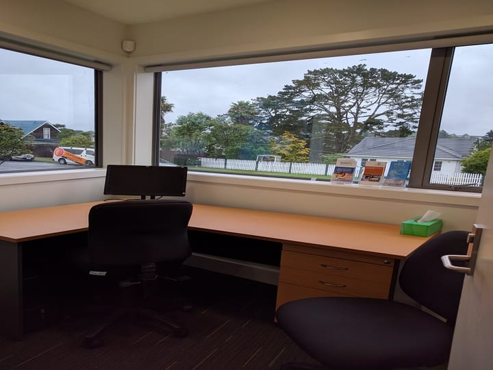Small office room with L-shaped desk, monitor, and panoramic windows to garden