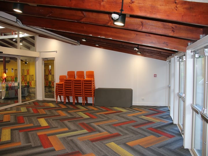 Bare hall with timber beam ceiling, herringbone carpet tiles, stacked orange chairs