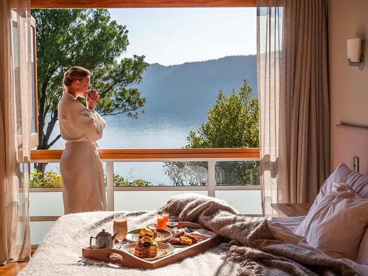 Bedroom with a water view, a person in a robe at the window, and a breakfast tray on the bed