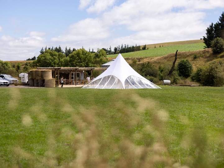 White peak tent on open farmland with rural hills behind, rustic barn building visible to side