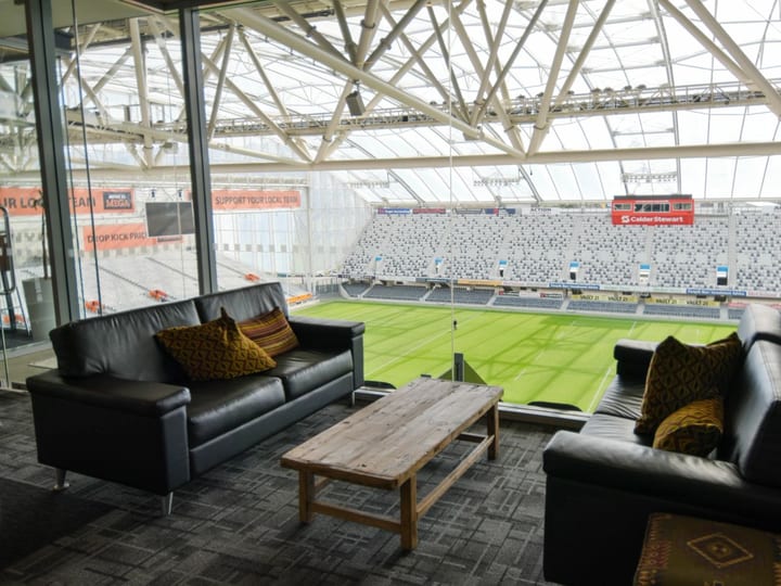 Stadium lounge with black leather sofas, timber coffee table, floor-to-ceiling windows overlooking playing field